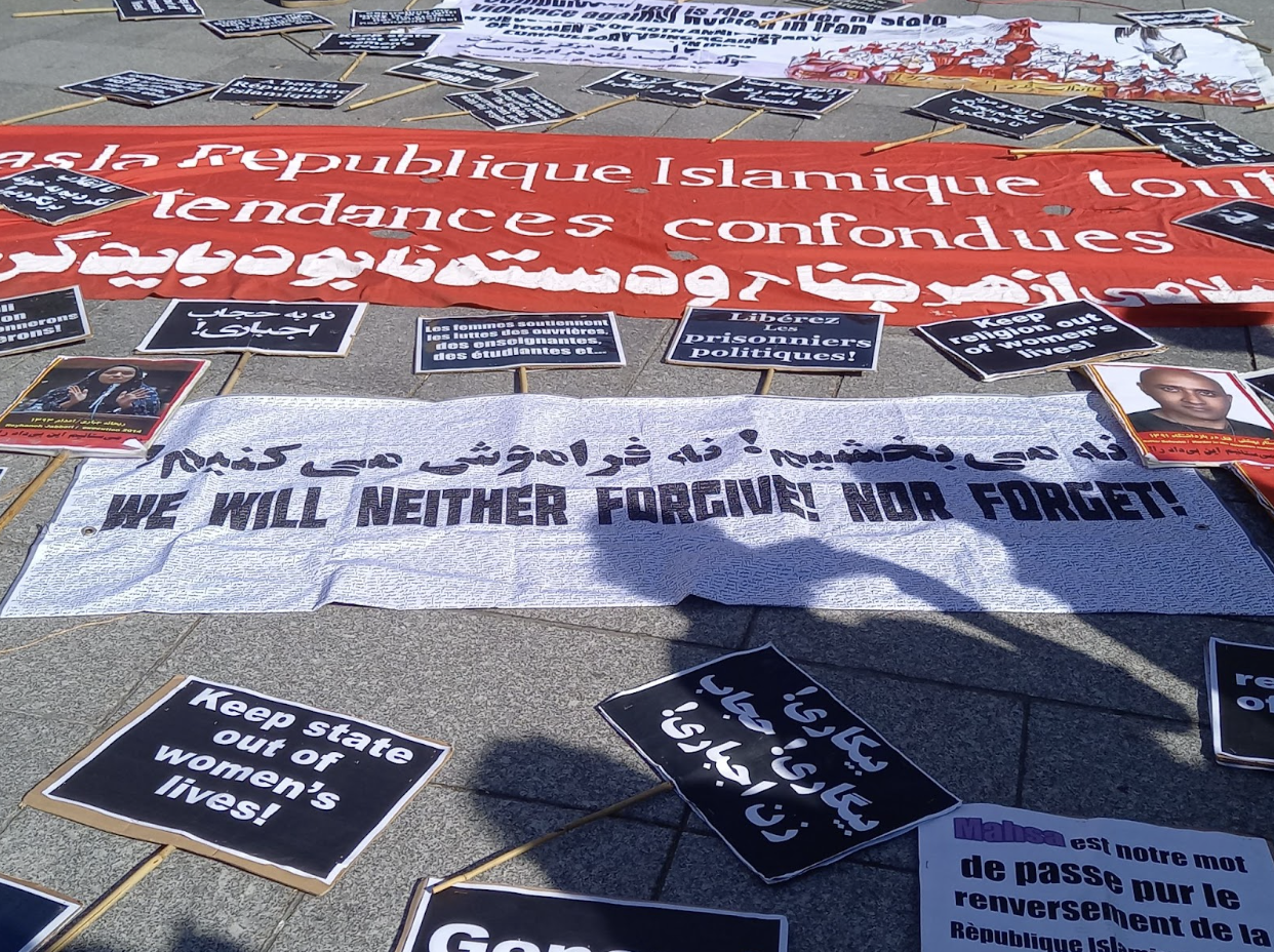 Des banderoles et des pancartes de plusieurs tailles sont posées sur le sol gris. Elles sont noires, blanches et rouges, avec des slogans écrits en anglais, français et persan.